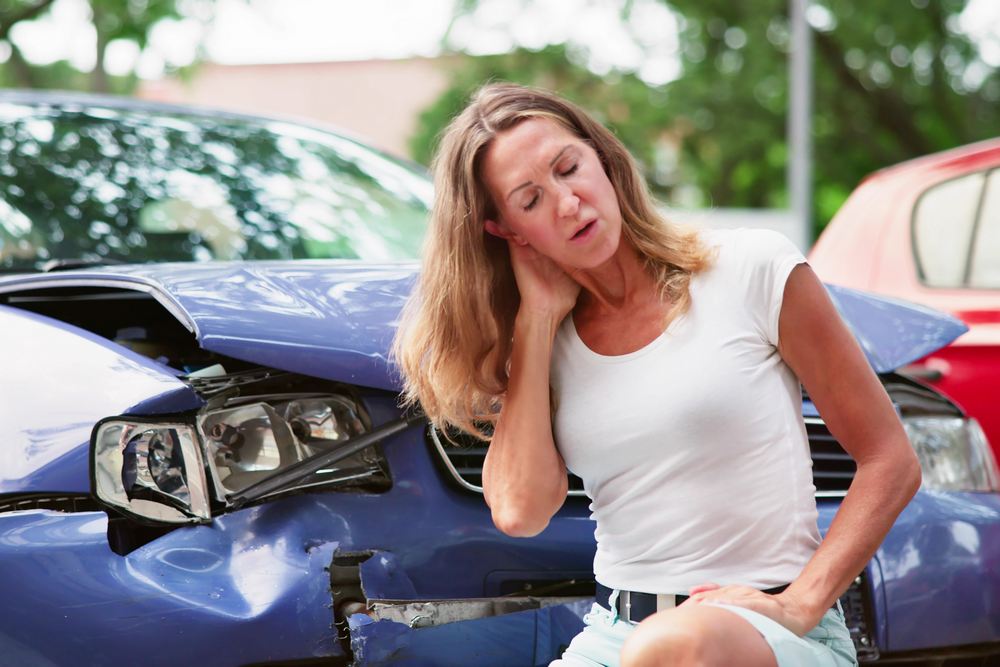 Advanced Health Solutions: woman holding her neck in discomfort beside a blue car with a crumpled front end and broken headlight