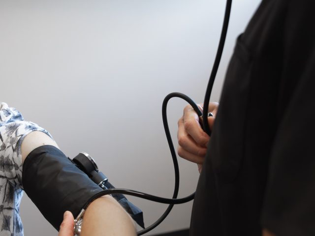 Advanced Health Solutions clinician using a stethoscope at a patient's inner elbow to take a blood pressure reading in an exam room