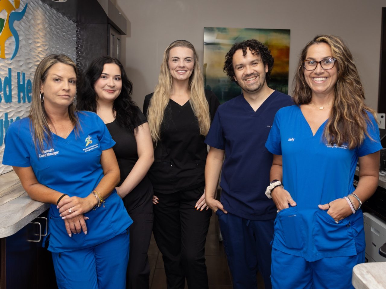 Advanced Health Solutions team of five in blue and black scrubs smiling together in the reception area beside the clinic's logo wall