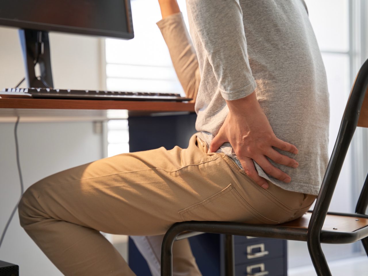 Advanced Health Solutions: person in a gray shirt and khaki pants holding their lower back while seated at a computer desk